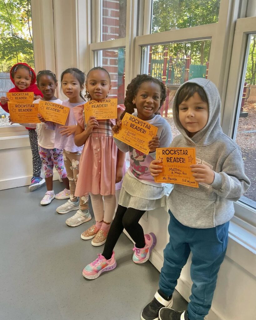 Diverse group of elementary students at Delphi Academy's Milton campus proudly displaying their 'Rockstar Reader' achievement certificates near a sunny classroom window